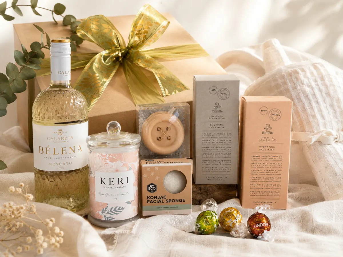 Gift box tied with a gold ribbon beside soft linen, moscato and body care